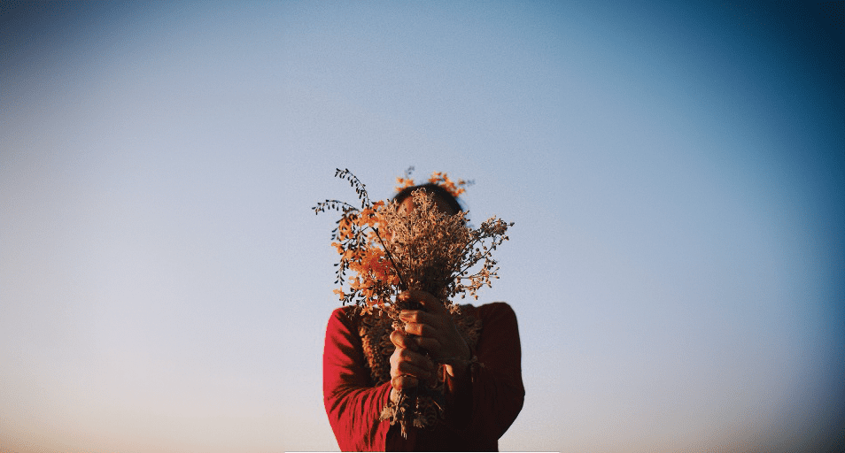 A person obscured behind a bouquet of wildflowers held up to their face, against a clear blue sky, symbolizing the uniqueness and varied perspectives of autistic individuals.